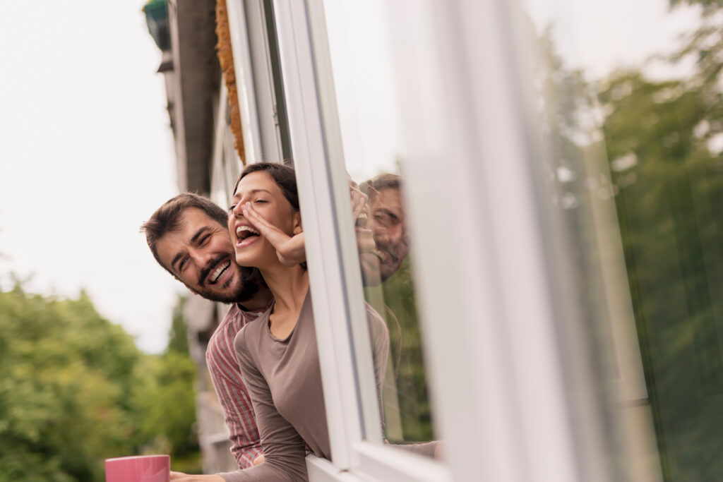 Couple leaning through an opened window, drinking coffee and calling friends passing by