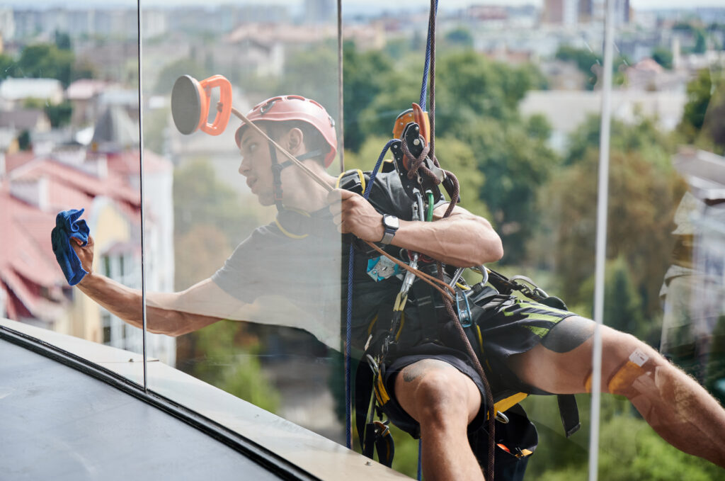 Industrial mountaineering cleaning service worker hanging on rope and wiping window. View from inside building. Cleaner using safety lifting equipment while cleaning glass of high-rise building.