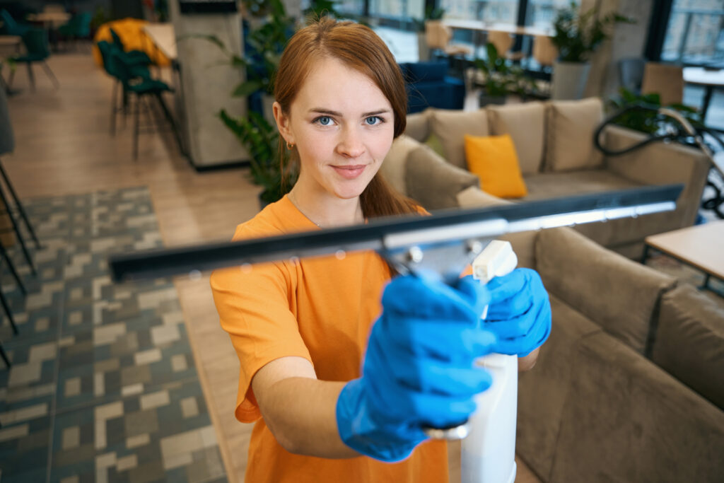 Red-haired cleaning lady in uniform and protective gloves uses glass scraper and spray to work